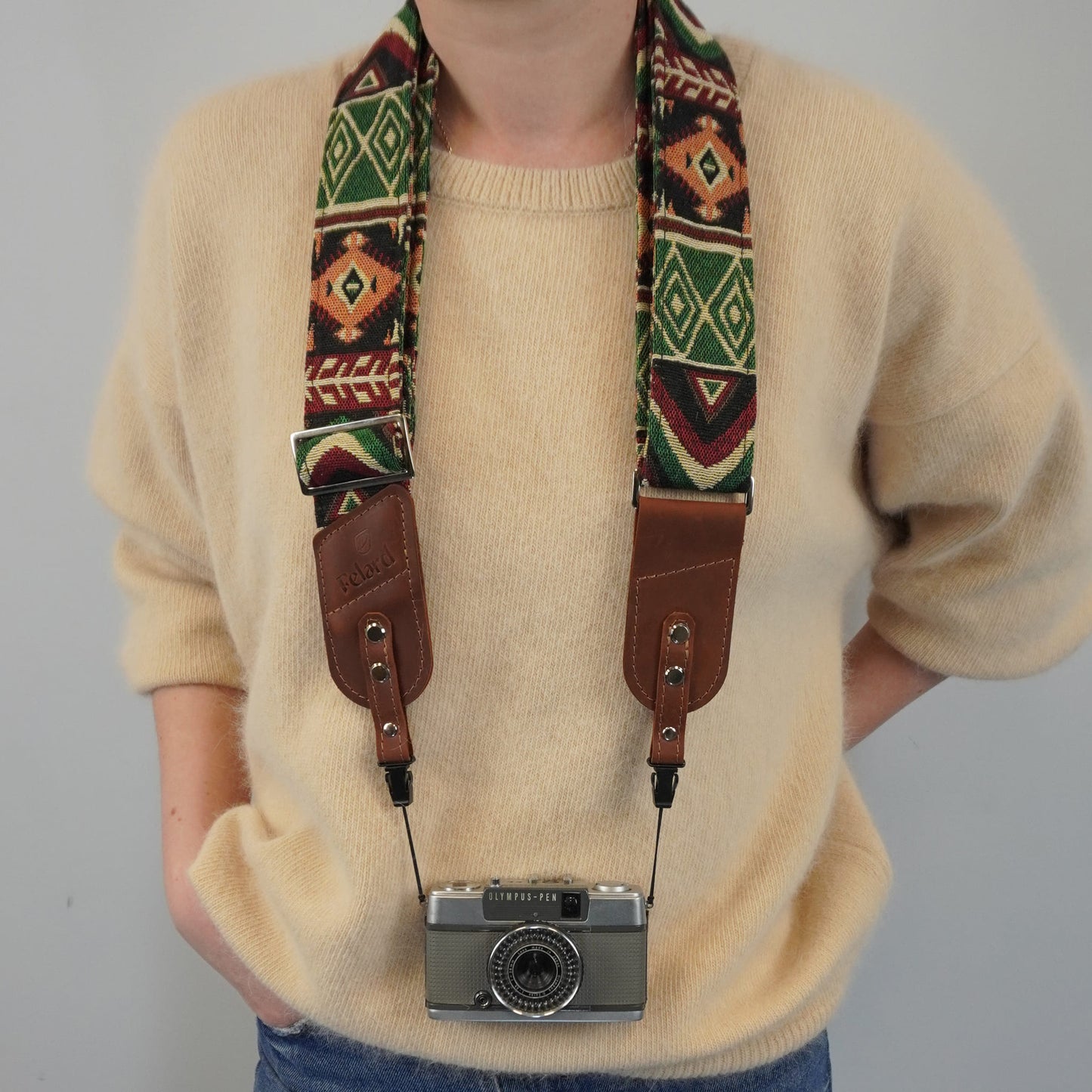 A person models a patterned camera strap with brown leather sections, holding a silver film camera against a plain light gray background.