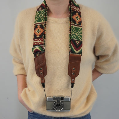 A person models a patterned camera strap with brown leather sections, holding a silver film camera against a plain light gray background.