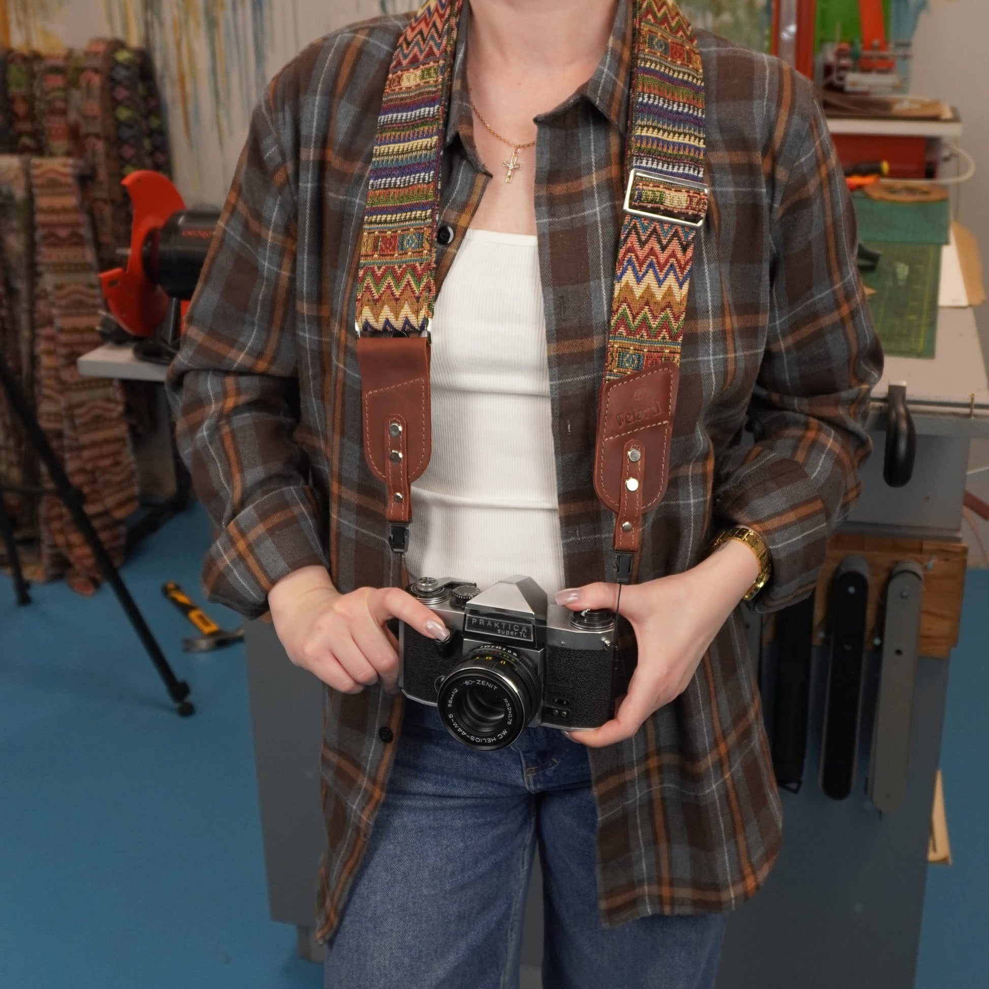 A person holding a vintage black camera with a colorful patterned camera strap. The background shows a workshop or studio environment with sewing machines and fabrics.