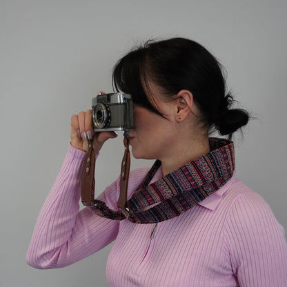 A woman wearing a colorful patterned neck camera strap with a brown leather strap, holding a vintage silver camera against her eye. The background is a plain light gray wall.