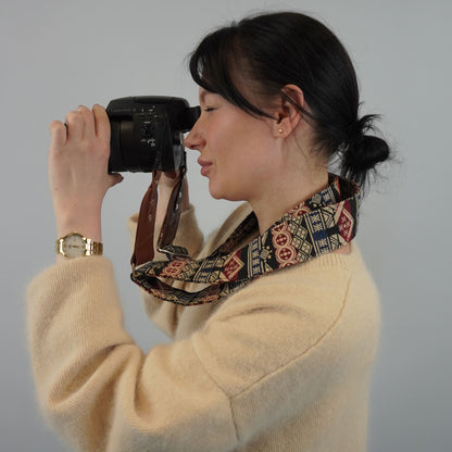 A woman with patterned camera strap around her neck, holding a black camera to her eye against a plain light gray background.