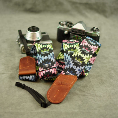 A set of camera straps with a colorful geometric pattern and brown leather ends, displayed alongside two vintage cameras on a textured gray surface.