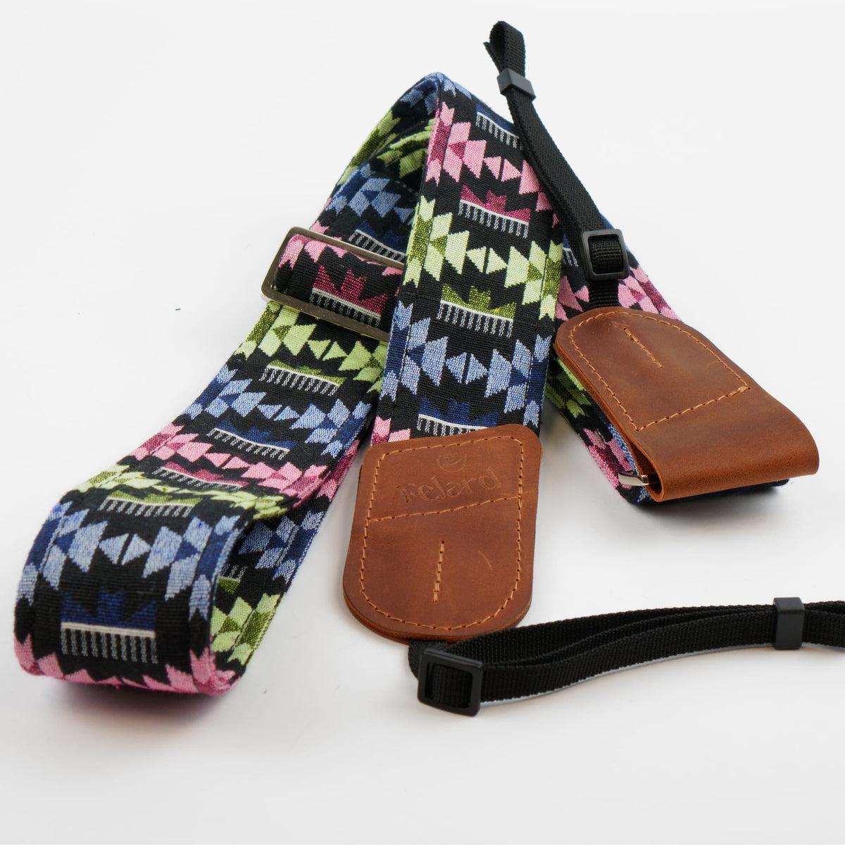 A camera strap with a colorful geometric pattern in pink, green, blue, and black, featuring brown leather end caps and a black adjustable webbing strap with a plastic buckle.