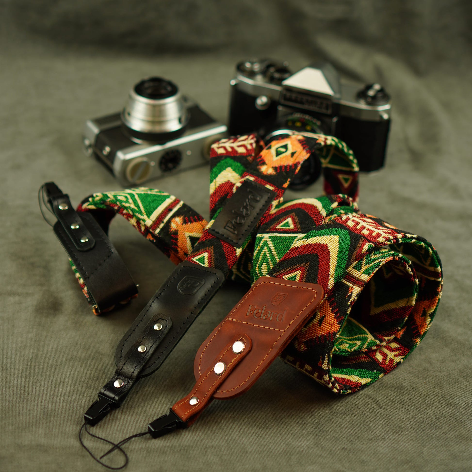 A rolled-up camera straps with a colorful geometric pattern in green, red, orange, and cream, combined with black leather and brown leather sections, displayed on a textured gray surface with vintage cameras in the background.