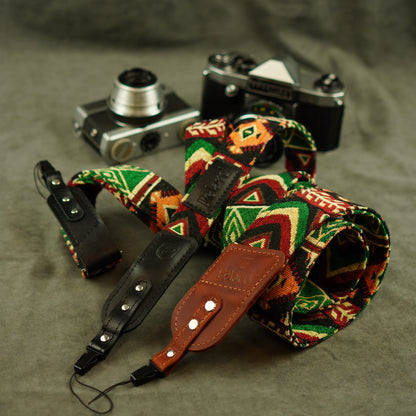 A rolled-up camera straps with a colorful geometric pattern in green, red, orange, and cream, combined with black leather and brown leather sections, displayed on a textured gray surface with vintage cameras in the background.