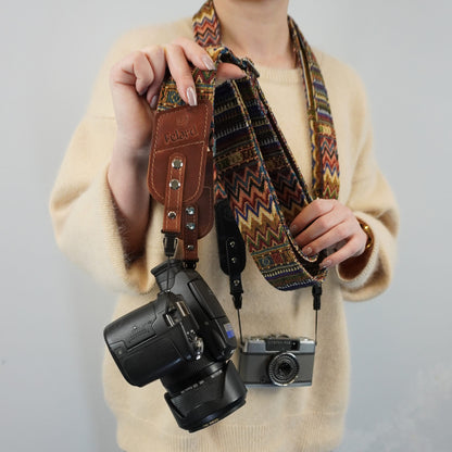 A person holds up a brown leather camera strap with silver rivets and a colorful geometric patterned camera strap. A black DSLR camera and a silver rangefinder are hanging from the straps against a plain light gray background.