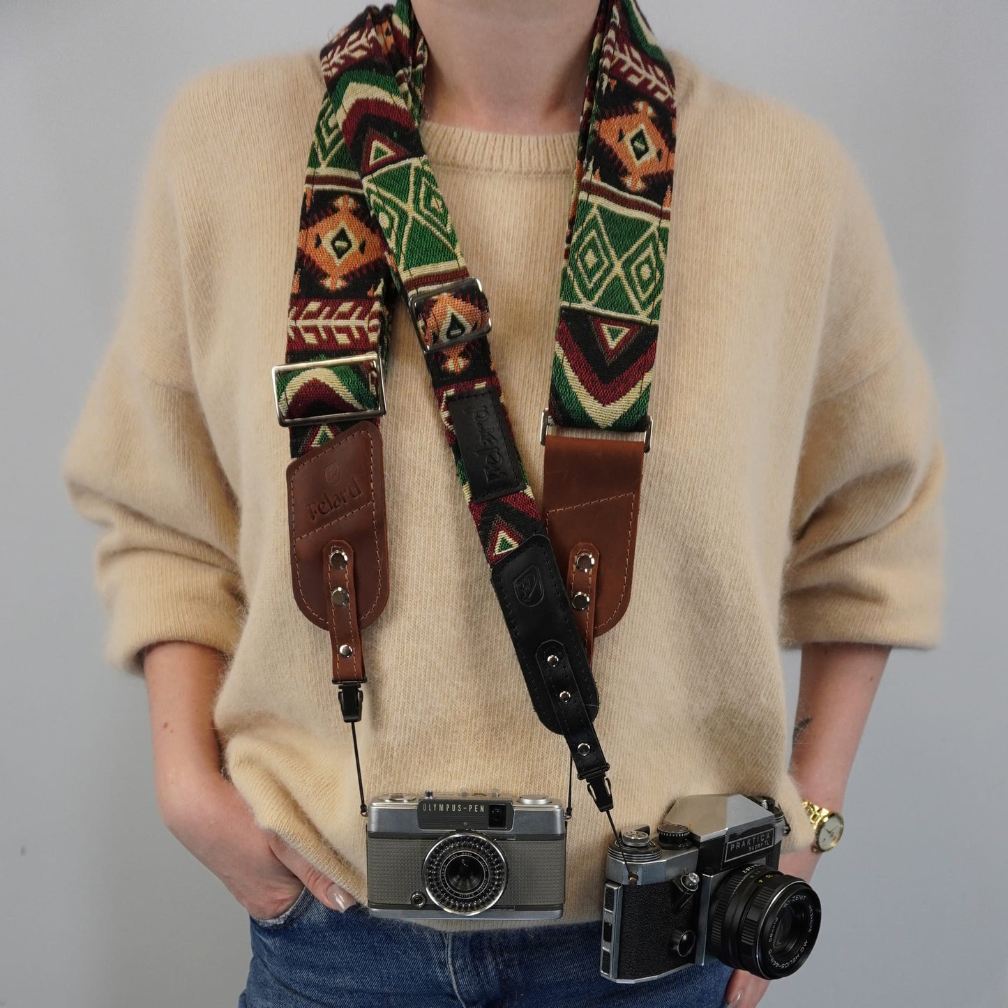 A person carries a patterned camera strap with brown leather sections over their shoulders, holding two vintage cameras.
