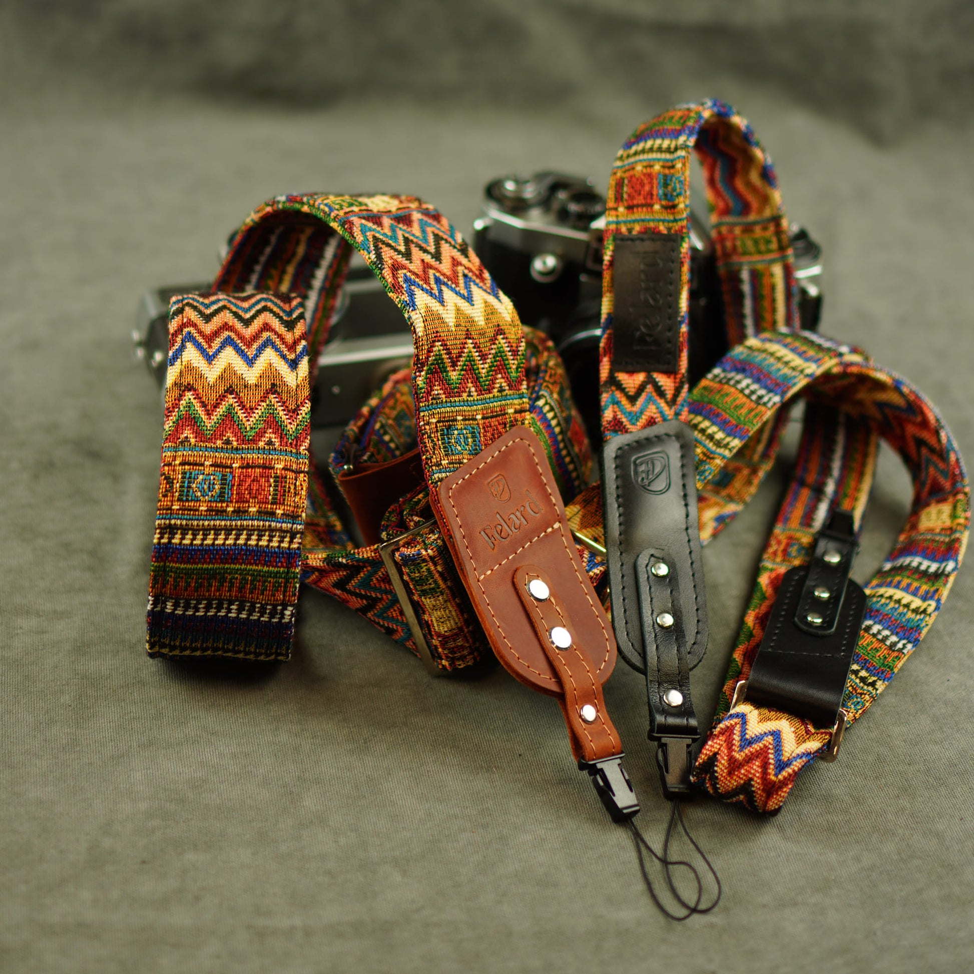 A set of camera straps with colorful geometric woven patterns and brown leather accents, displayed on a textured gray surface next to a vintage camera.