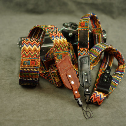 A set of camera straps with colorful geometric woven patterns and brown leather accents, displayed on a textured gray surface next to a vintage camera.