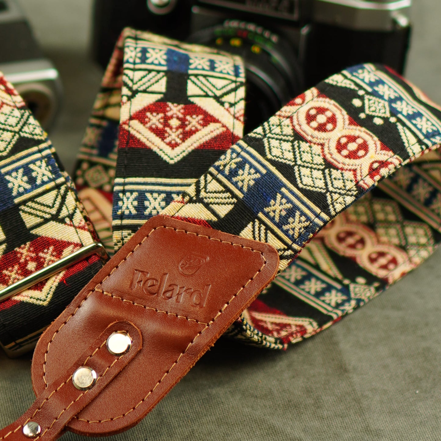 A close-up of a patterned camera strap with a brown leather. The strap features a multi-colored geometric and Nordic-style pattern in red, blue, beige, and black.