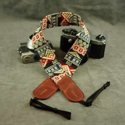 A patterned camera strap with brown leather ends and black adjustable ties, displayed in front of two vintage cameras on a gray textured surface.