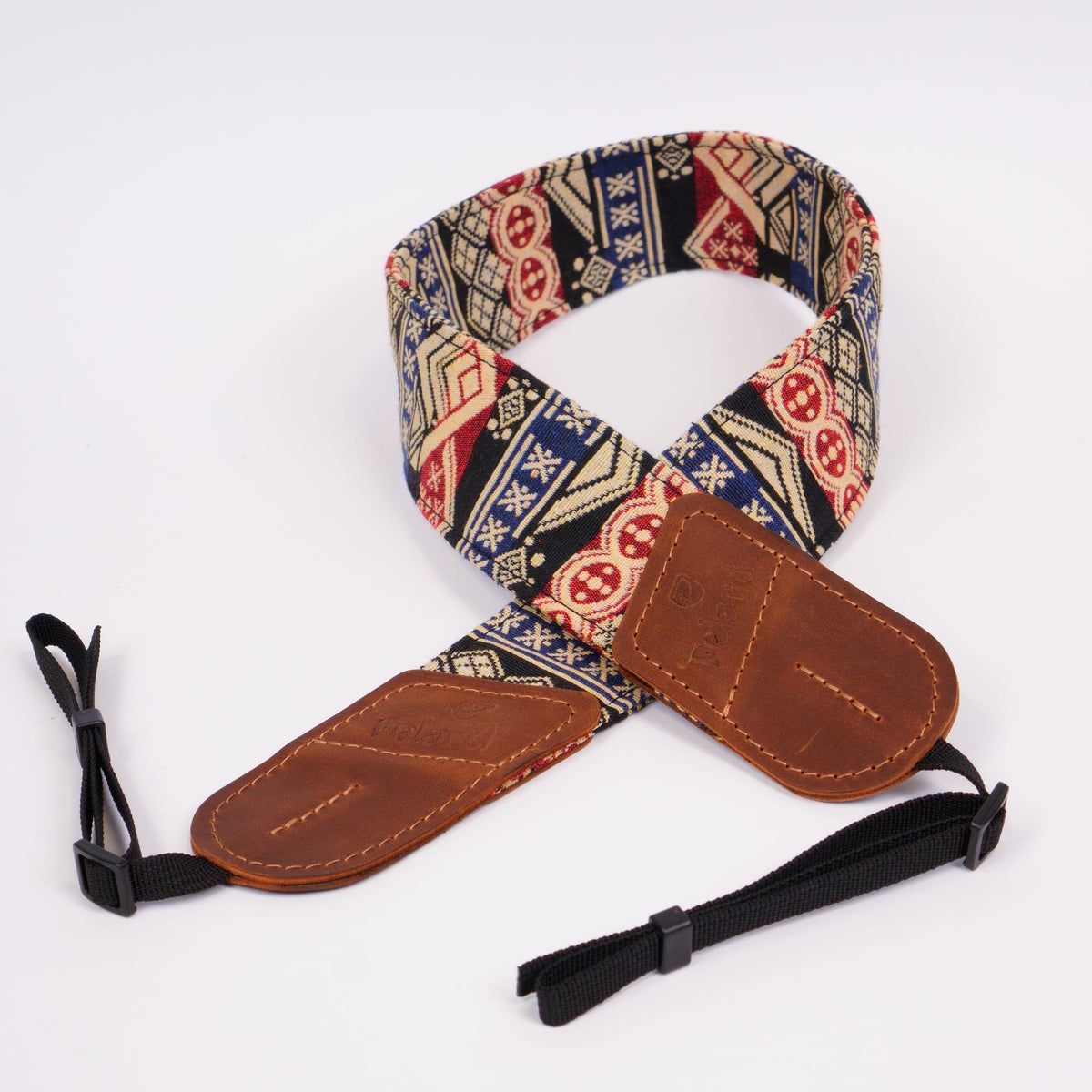 A fabric camera strap with a colorful geometric pattern in navy, red, beige, and white, featuring brown leather ends and black webbing loops.
