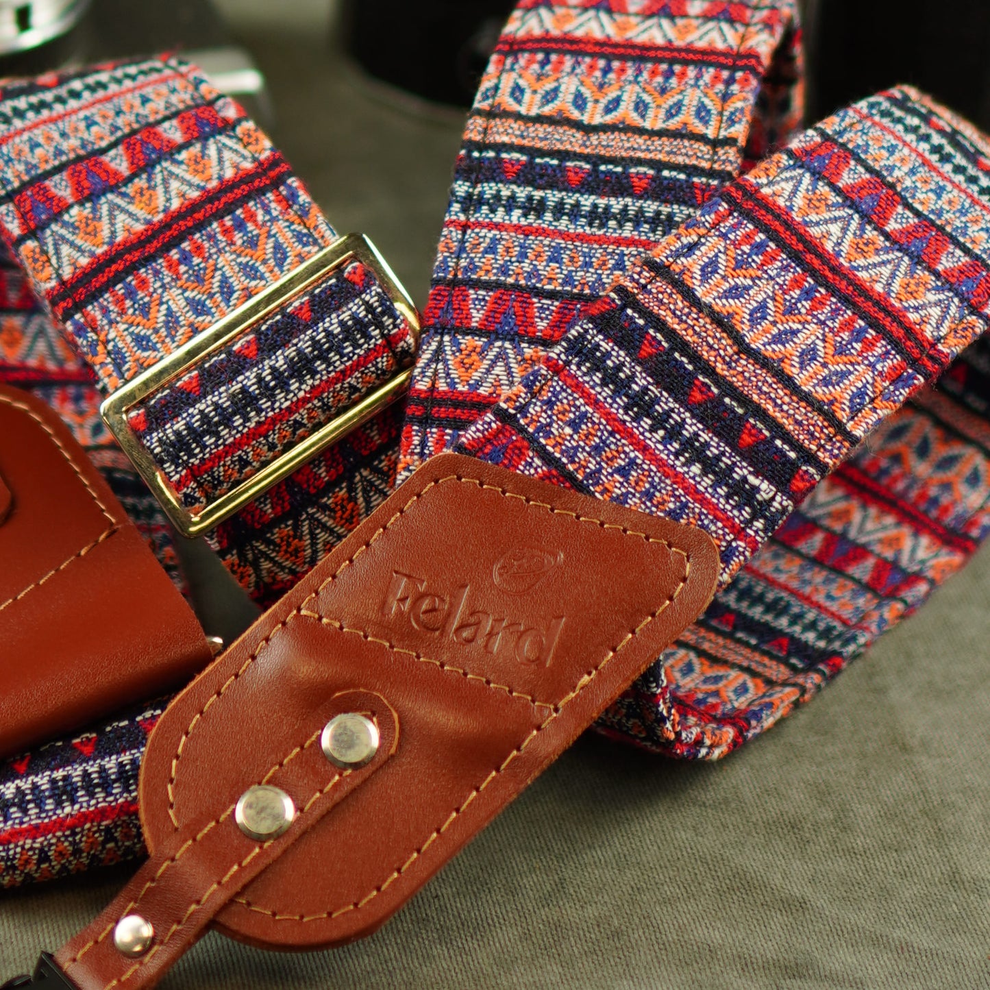 A close-up of a woven belt with a colorful geometric pattern in red, blue, orange, and white, featuring a brown leather keeper and gold-tone buckle.