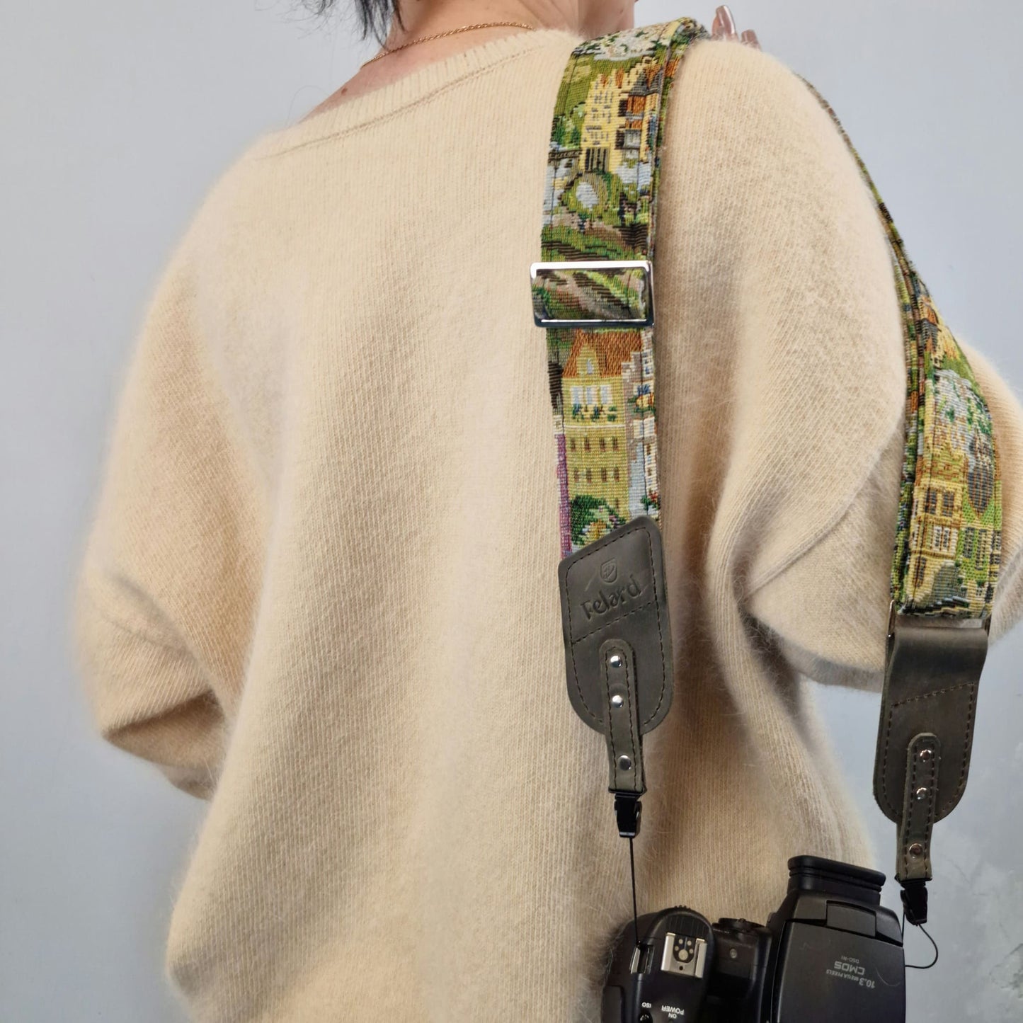 A close-up of a person from behind wearing a cream-colored knit sweater and carrying a camera with a colorful printed camera strap. The strap features a scenic architectural pattern in green, yellow, and beige tones, with gray leather hardware and a silver metal buckle.