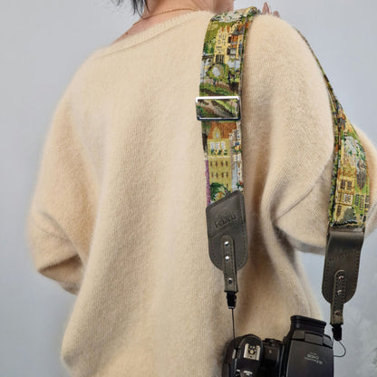 A close-up of a person from behind wearing a cream-colored knit sweater and carrying a camera with a colorful printed camera strap. The strap features a scenic architectural pattern in green, yellow, and beige tones, with gray leather hardware and a silver metal buckle.