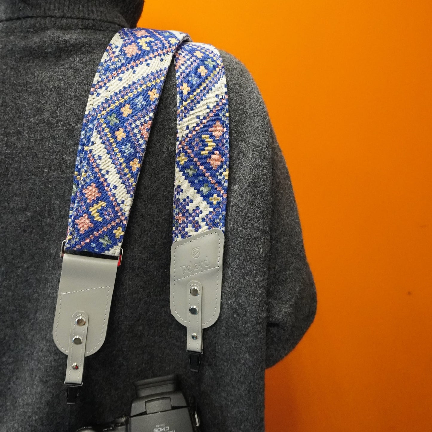 A close-up of a camera strap with a blue, white, and pink geometric pattern, worn over the shoulder of a person wearing a dark gray sweater against a solid orange background.