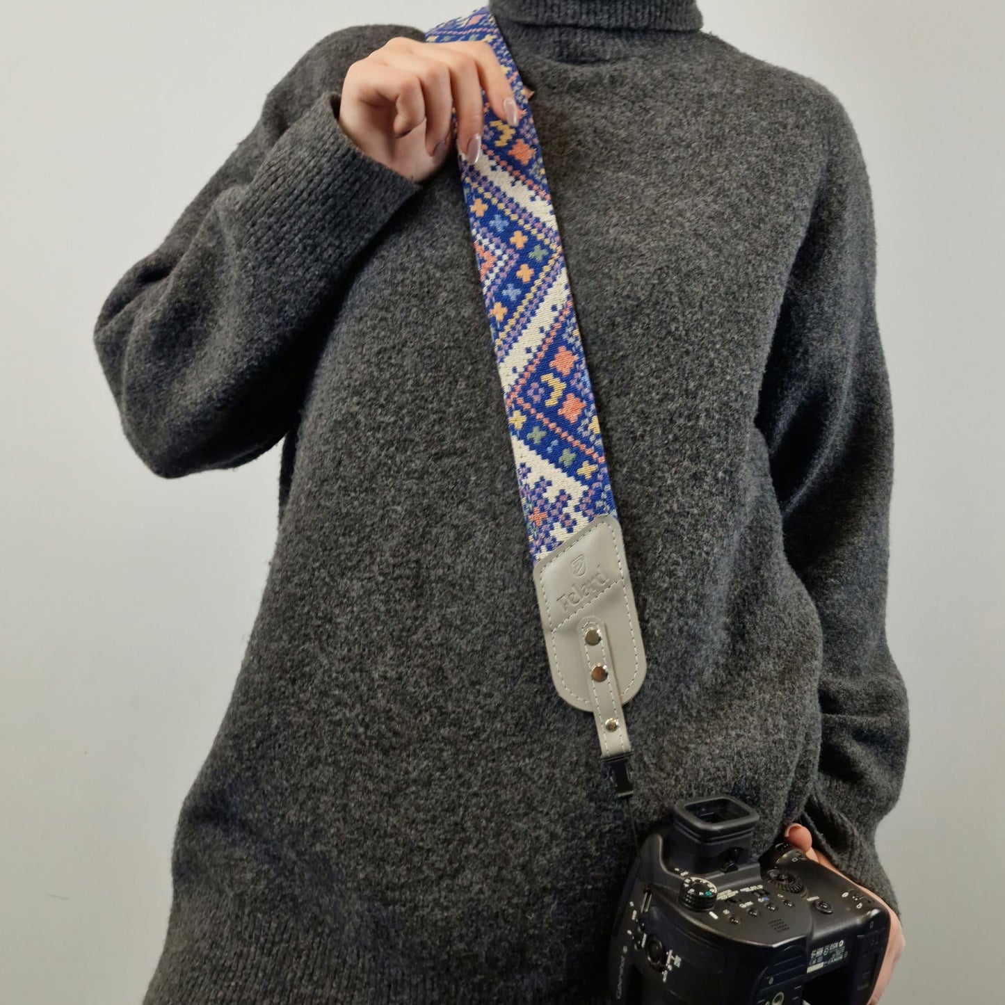 A person carries a camera with a patterned camera strap. The strap features a blue, white, and orange geometric design with a light gray leather end piece.