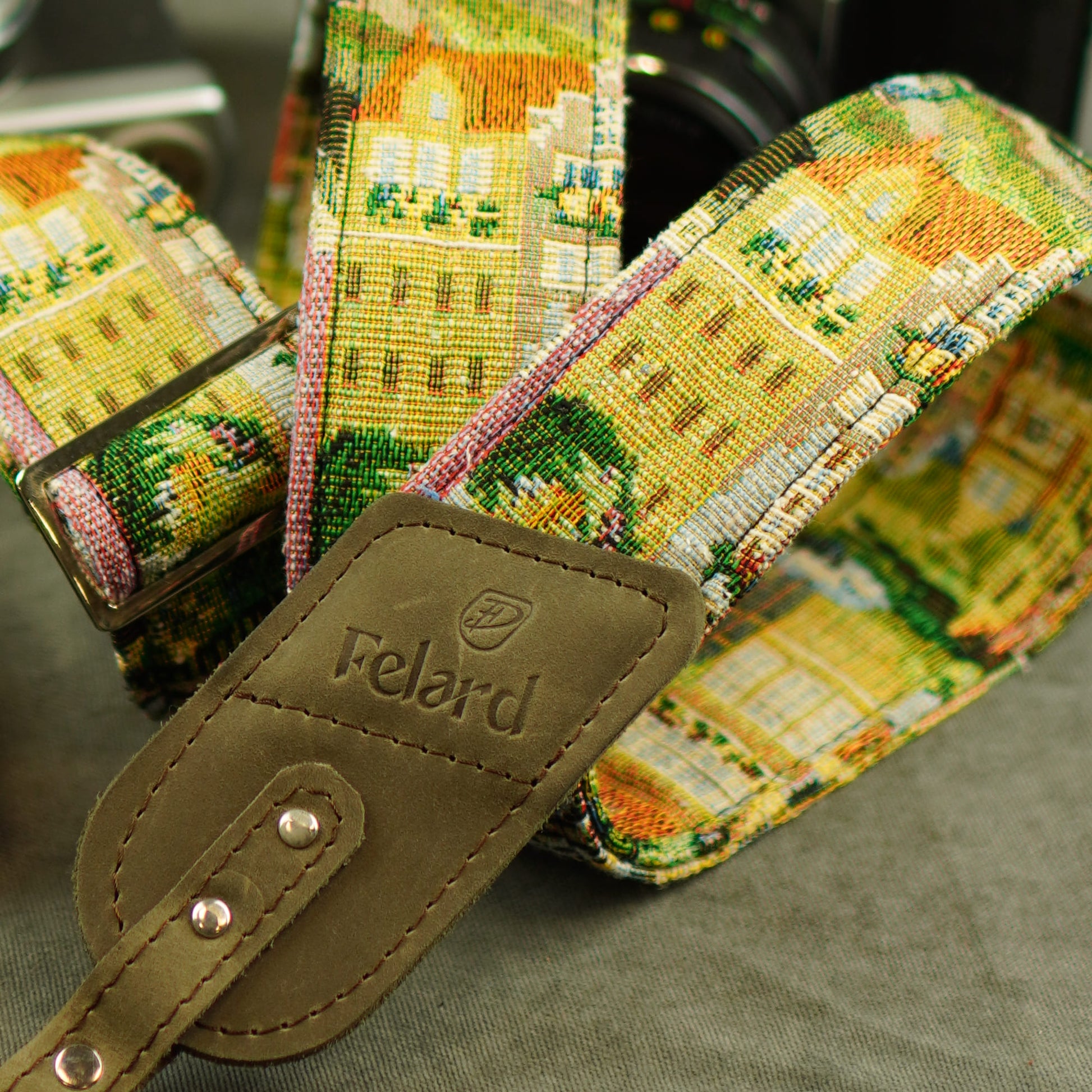 A close-up of a woven camera strap featuring a colorful architectural village pattern in yellow, green, pink, and white, with a brown leather brand tab stamped 'Felard'.