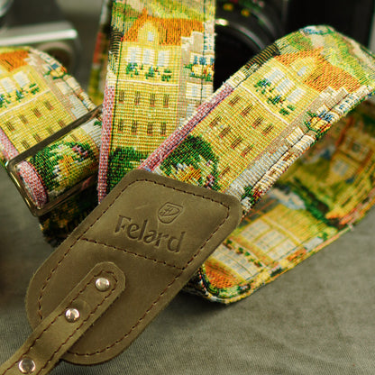A close-up of a woven camera strap featuring a colorful architectural village pattern in yellow, green, pink, and white, with a brown leather brand tab stamped 'Felard'.