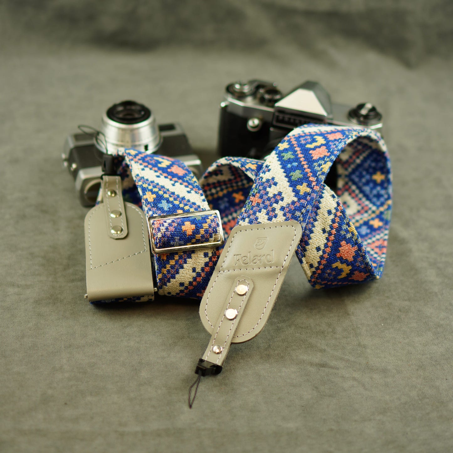 A woven camera strap with a blue, beige, and orange geometric pattern, attached to light beige leather keeper, displayed on a textured gray surface next to vintage cameras.