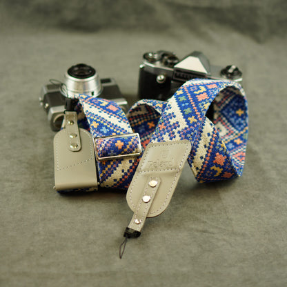 A woven camera strap with a blue, beige, and orange geometric pattern, attached to light beige leather keeper, displayed on a textured gray surface next to vintage cameras.