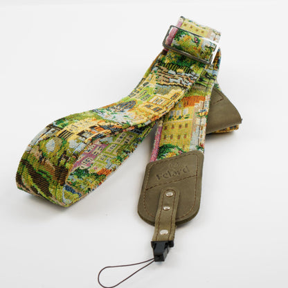 A fabric camera strap featuring a colorful, detailed landscape tapestry pattern with yellow buildings and green trees, accented by an olive green leather patch embossed with the brand 'Felard'.