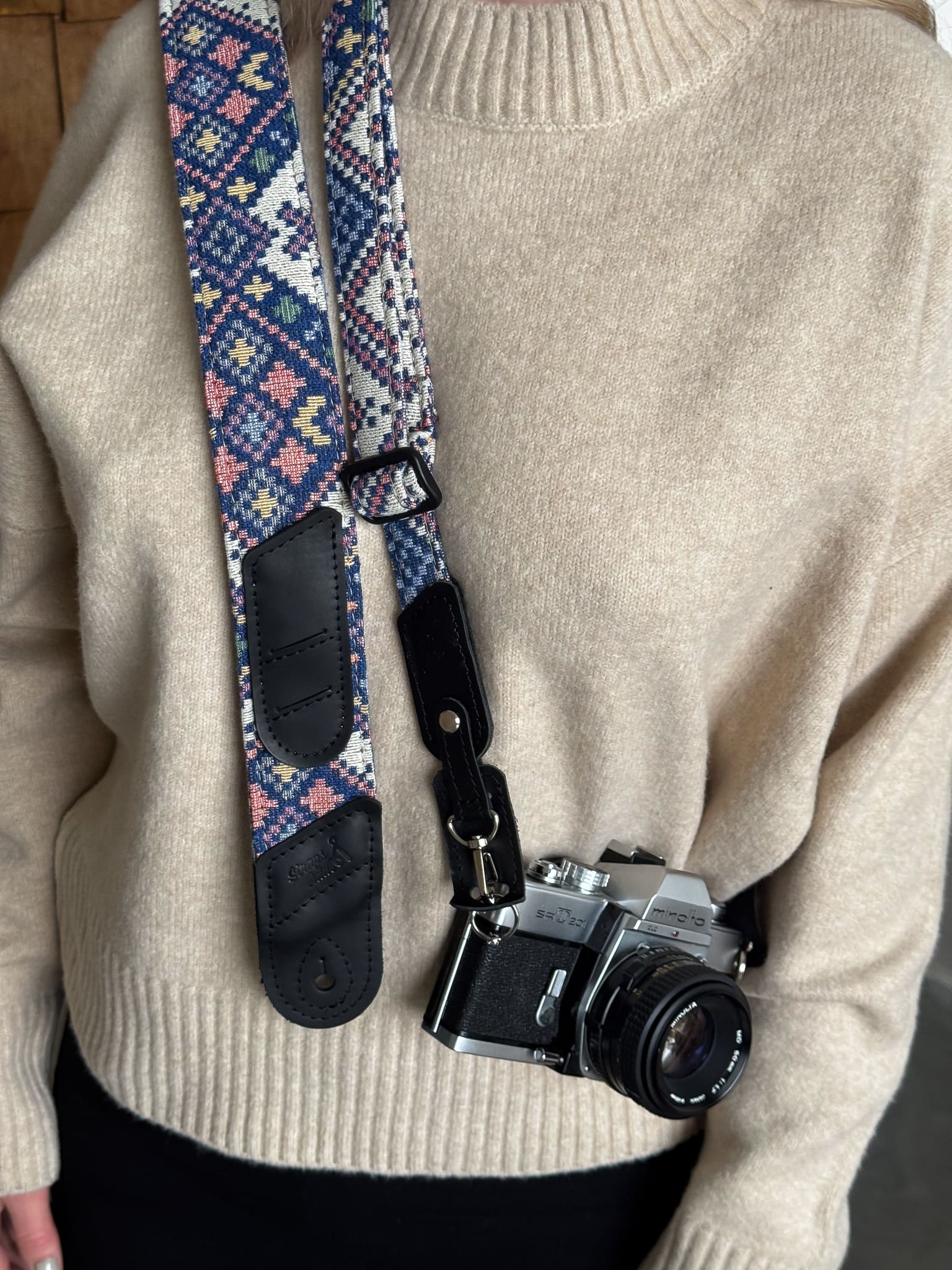 A close-up of a person carrying a vintage silver and black camera with a patterned woven strap. The strap features a blue, pink, and white geometric design with black leather ends.