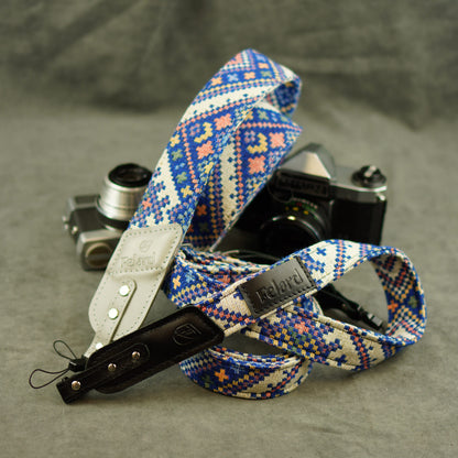 A patterned camera strap with blue, pink, and cream geometric embroidery, shown draped over two cameras on a gray textured background. The strap features a light beige leather section and a black leather keeper.