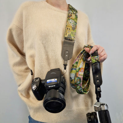 A person wearing a beige sweater models a colorful illustrated camera strap with gray metal hardware, holding a Sony DSLR camera and another camera in one hand.