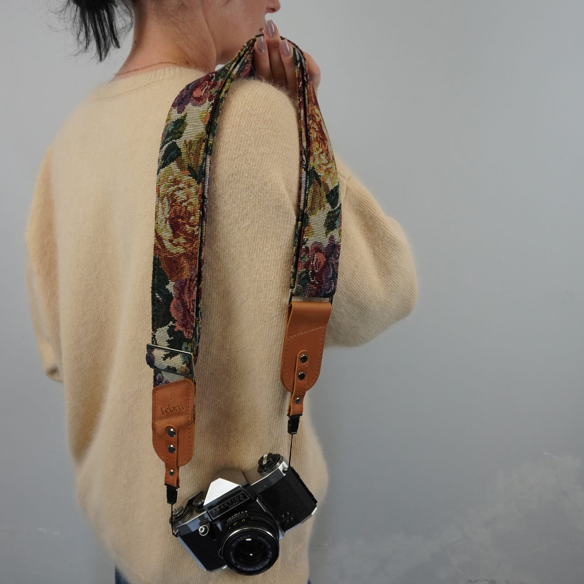 A person holds a floral camera strap with tan leather ends, carrying a black film camera over their shoulder against a plain gray wall.