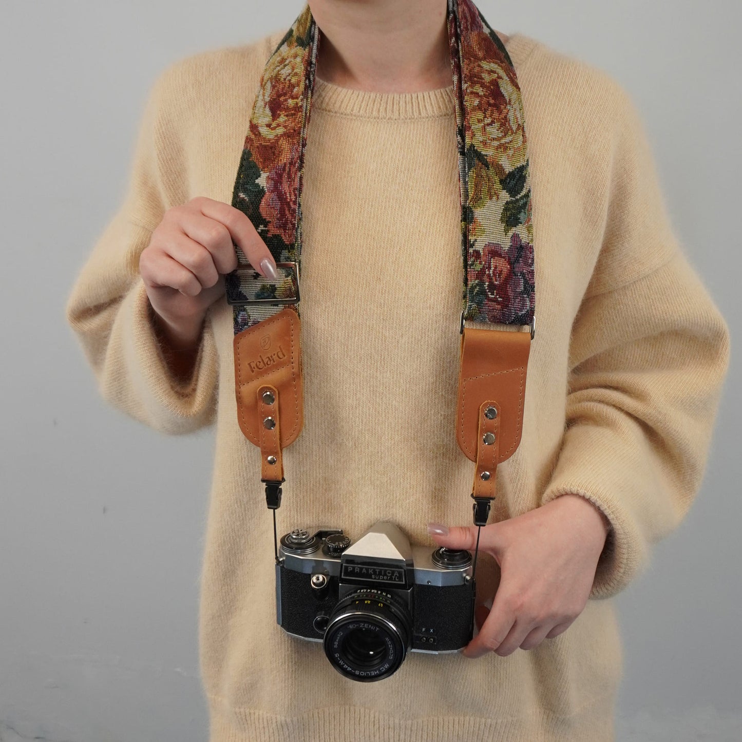 A person holds a vintage camera with a floral-patterned camera strap featuring tan leather accents, against a plain light gray background.