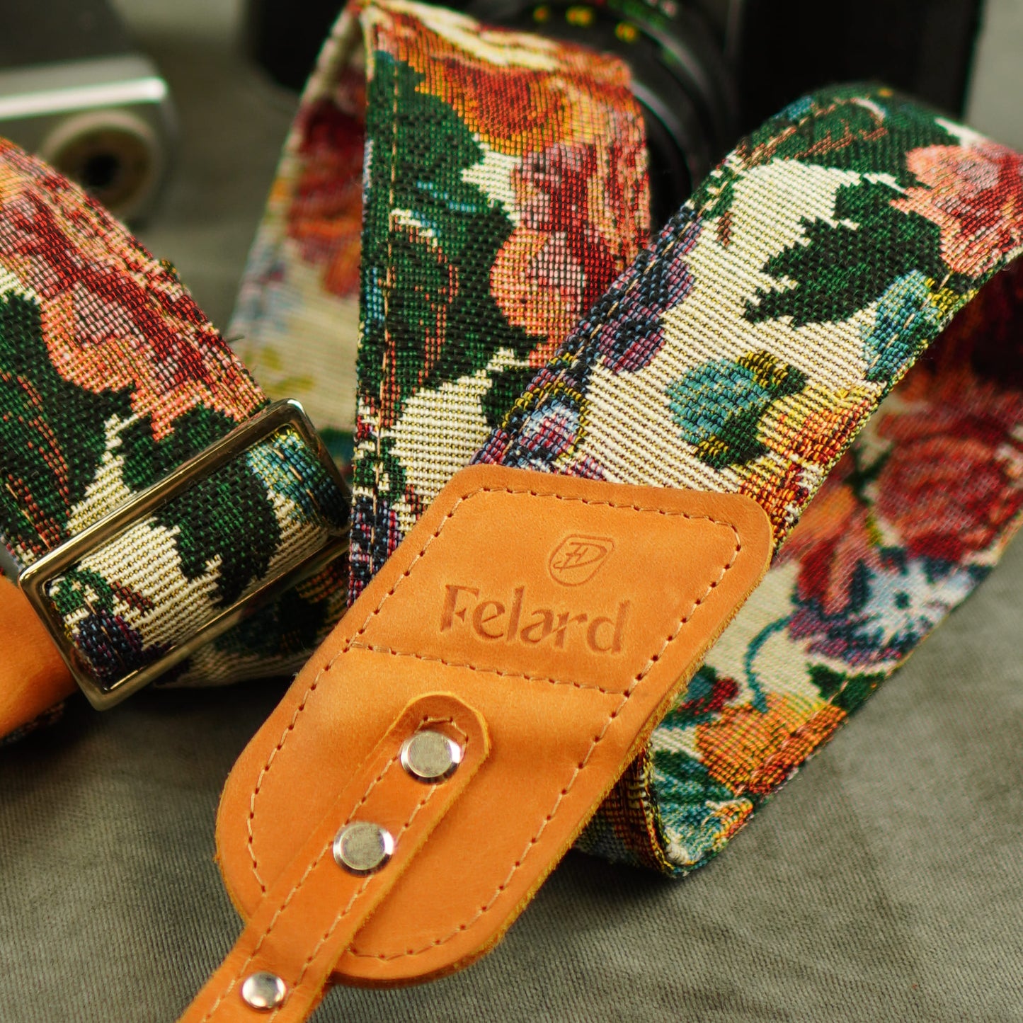 A close-up of a Felard camera strap featuring a colorful floral tapestry pattern in green, orange, red, and cream, with a tan leather buckle and logo patch.