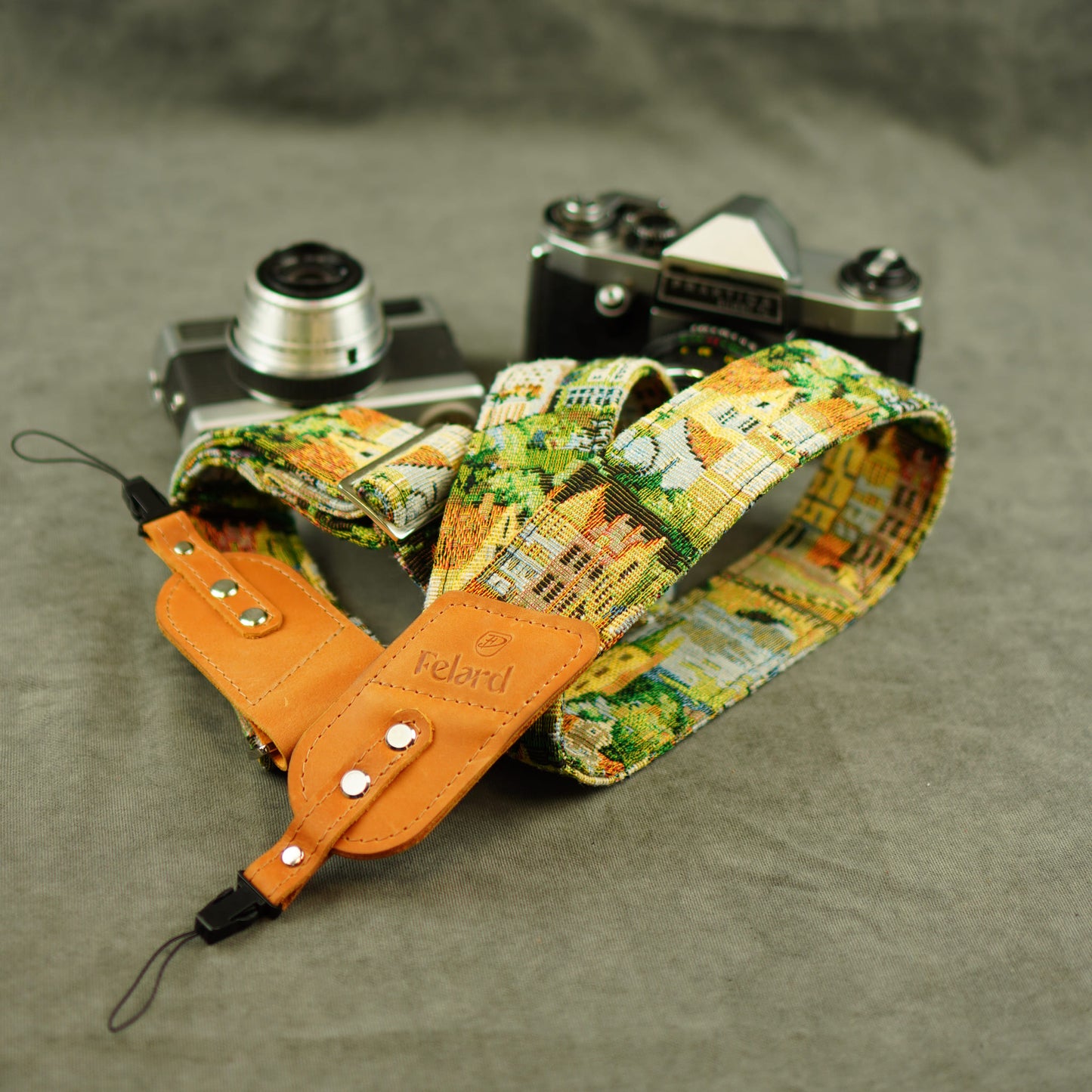 A fabric camera strap with a colorful cityscape print and tan leather, displayed on a gray textured surface with two vintage cameras behind it.