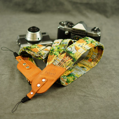 A fabric camera strap with a colorful cityscape print and tan leather, displayed on a gray textured surface with two vintage cameras behind it.