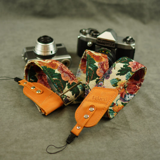A floral camera strap with tan leather accents displayed in front of two cameras on a textured gray surface. The strap features a multicolor floral pattern with green, red, and orange tones, and a tan leather section with metal rivets and a visible 'Felard' brand stamp.