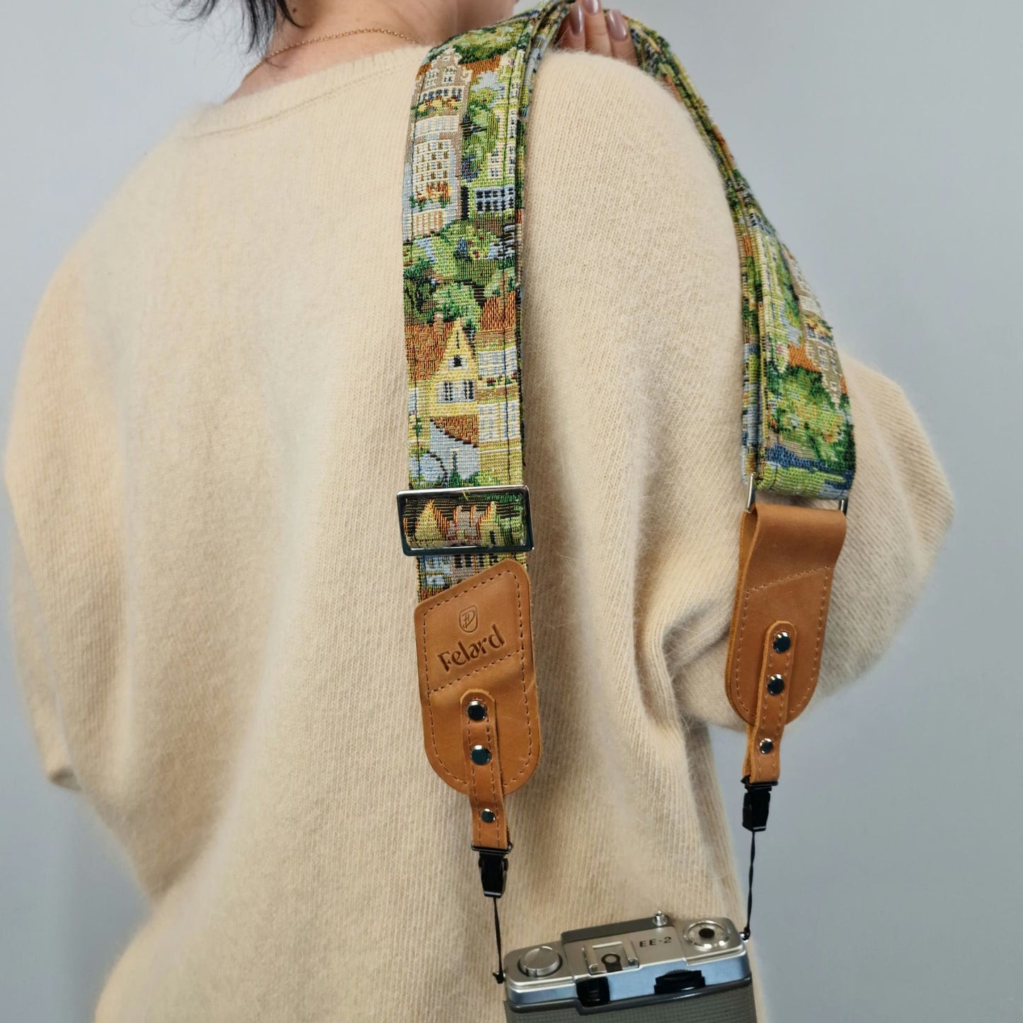 A close-up of a person from behind wearing a cream-colored knit sweater and carrying a Felard camera strap with a colorful village print and tan leather ends, holding a camera.