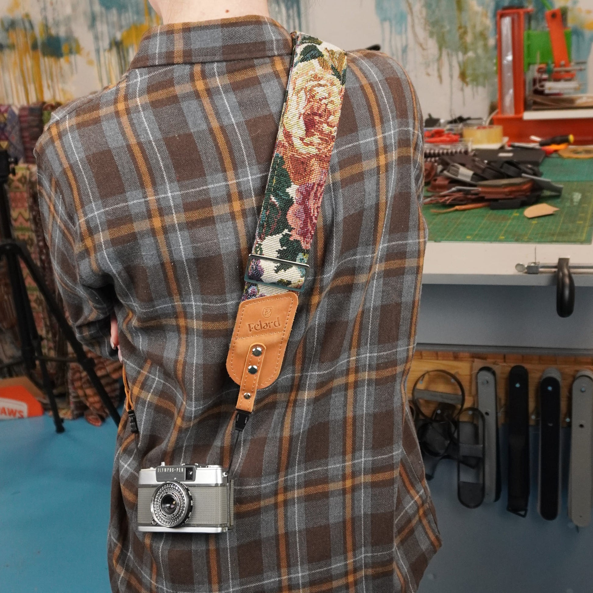A person viewed from behind, with a floral-patterned camera strap draped over the shoulder and a small silver camera hanging from the strap. The setting appears to be a workshop or studio with tools and materials in the background.