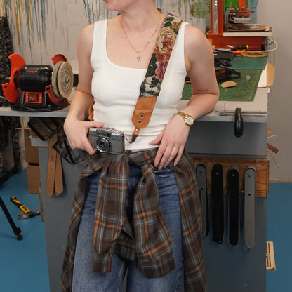 A person stands in a workshop setting, holding a camera and wearing a brown leather camera strap with a floral pattern and a tan logo patch. A plaid shirt is tied around the waist.
