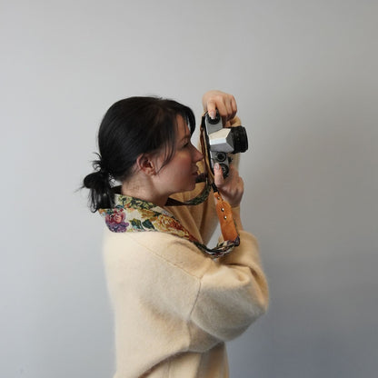 A woman holding a vintage camera with a floral-patterned strap, set against a plain light gray background.