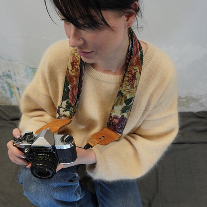 A person holds a silver film camera with a floral-patterned leather camera strap. The background shows a textured wall and a dark gray couch.
