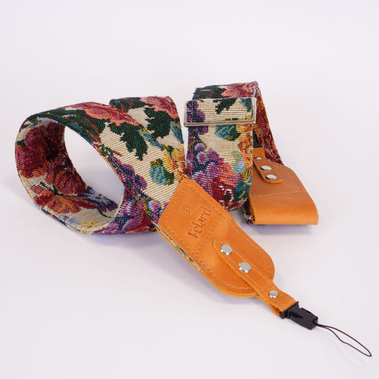 A wide camera strap featuring a colorful floral tapestry pattern and a tan leather ends, with a metal buckle and a small leather tag embossed with 'Felard'.