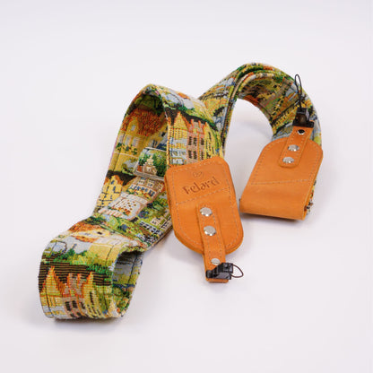 A camera strap featuring a colorful landscape patterned fabric and tan leather accents, displayed on a clean white background.