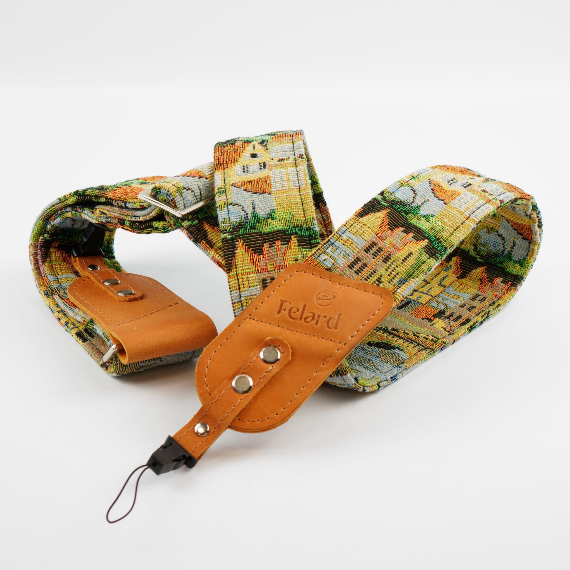 A fabric camera strap featuring a colorful architectural village print, with tan leather accents and a metal clip, shown on a light gray background.