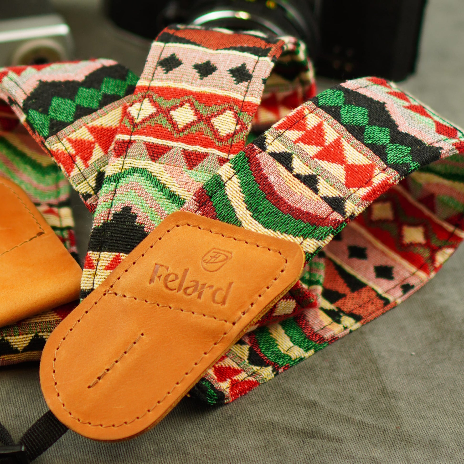 A close-up of a patterned camera strap with a tan leather Felard, featuring a geometric multicolor design in red, green, black, cream, and pink.