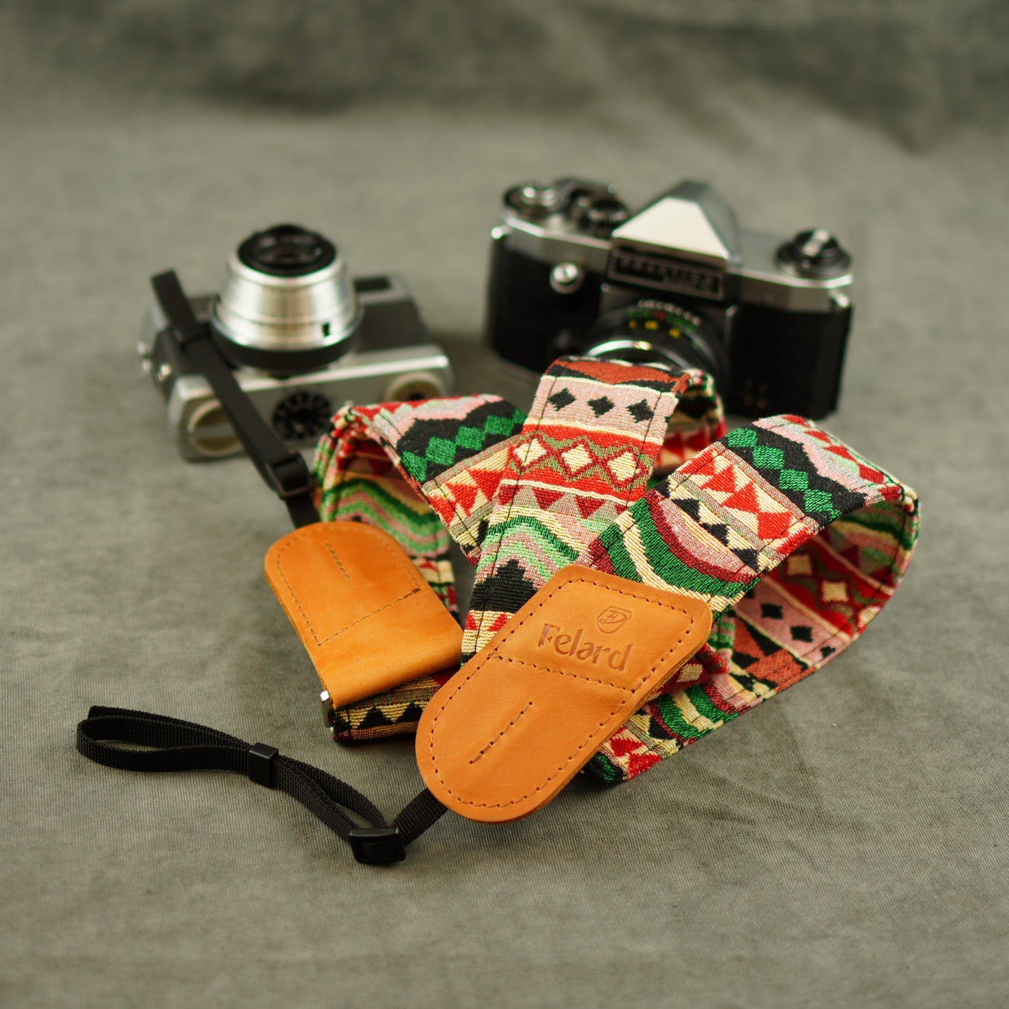 A patterned camera strap with tan leather displayed on a gray textured surface with two vintage cameras in the background. The leather loops are embossed with the brand 'Felard'.
