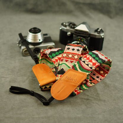 A patterned camera strap with tan leather displayed on a gray textured surface with two vintage cameras in the background. The leather loops are embossed with the brand 'Felard'.