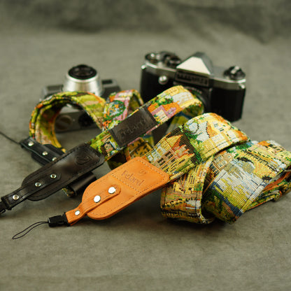 A vintage camera strap with a colorful cityscape print, brown leather attachments displayed on a gray textured surface next to two vintage cameras.