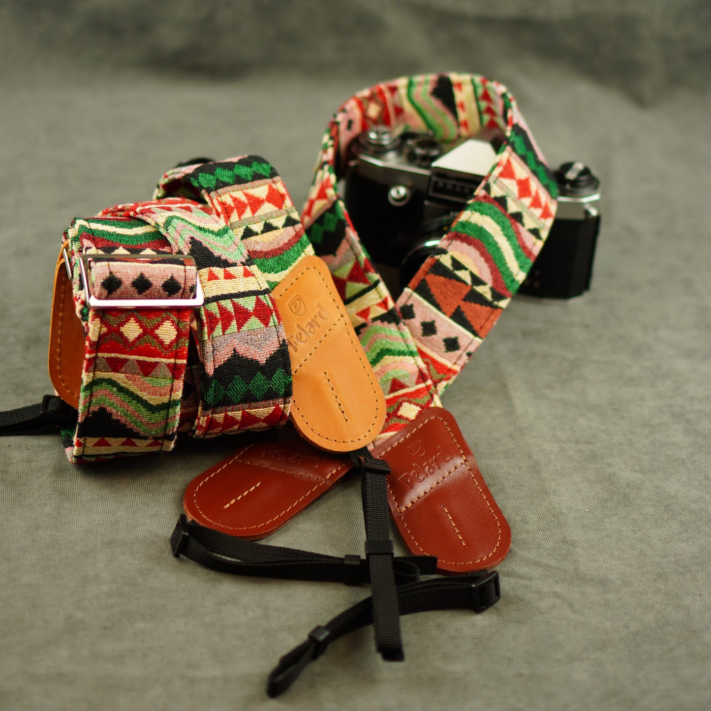 A close-up of a camera strap with a Southwestern geometric pattern in red, green, black, and beige, featuring tan leather accents, displayed on a gray textured surface with a vintage camera behind it.
