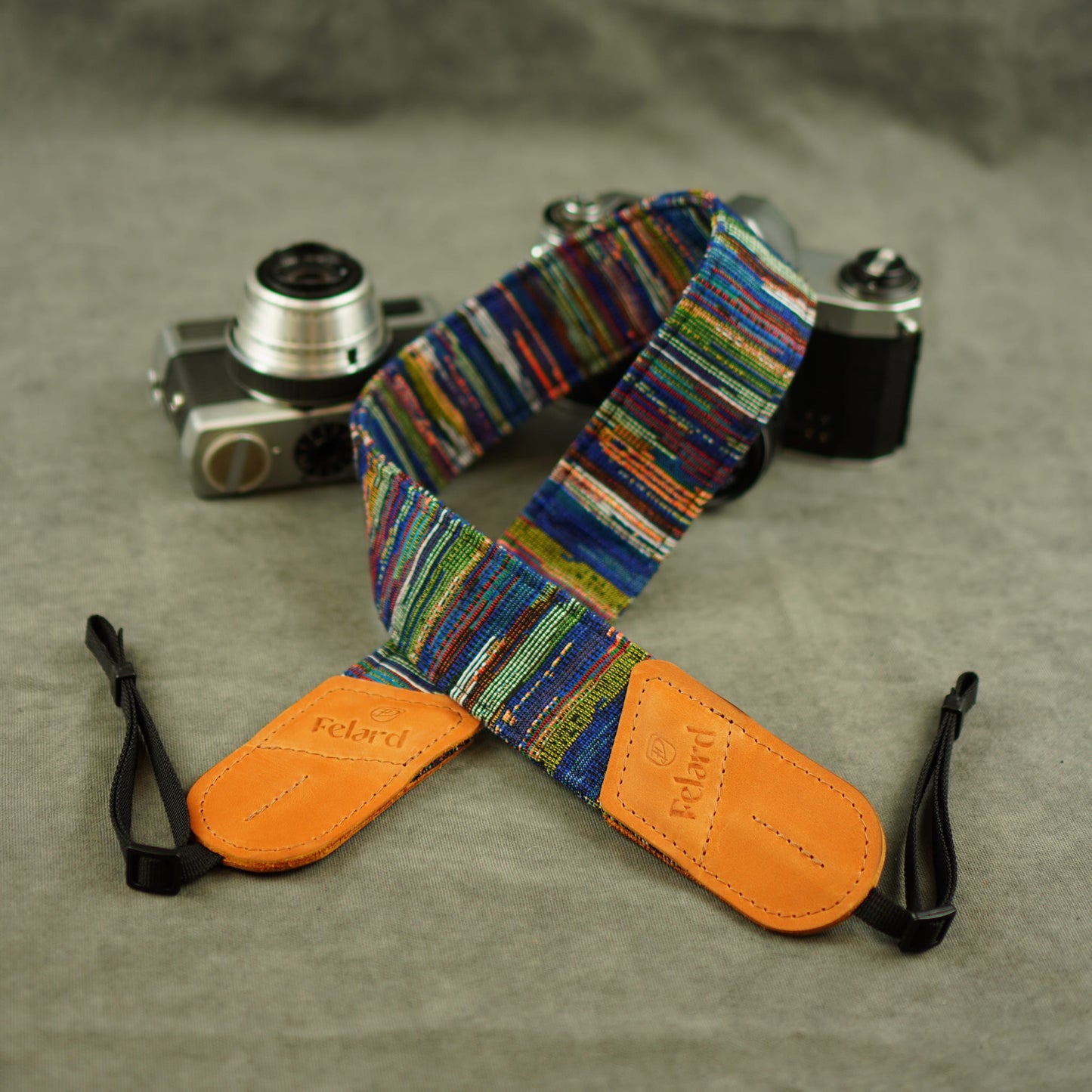 A colorful striped camera strap with tan leather end laid on a gray textured surface, with two cameras in the background. The strap has embossed 'Felard' branding on the tan end pieces.