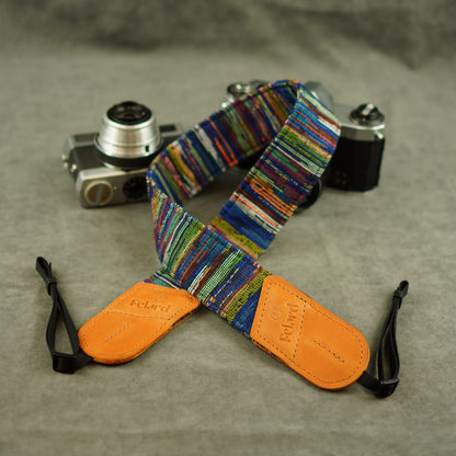 A colorful striped camera strap with tan leather end laid on a gray textured surface, with two cameras in the background. The strap has embossed 'Felard' branding on the tan end pieces.